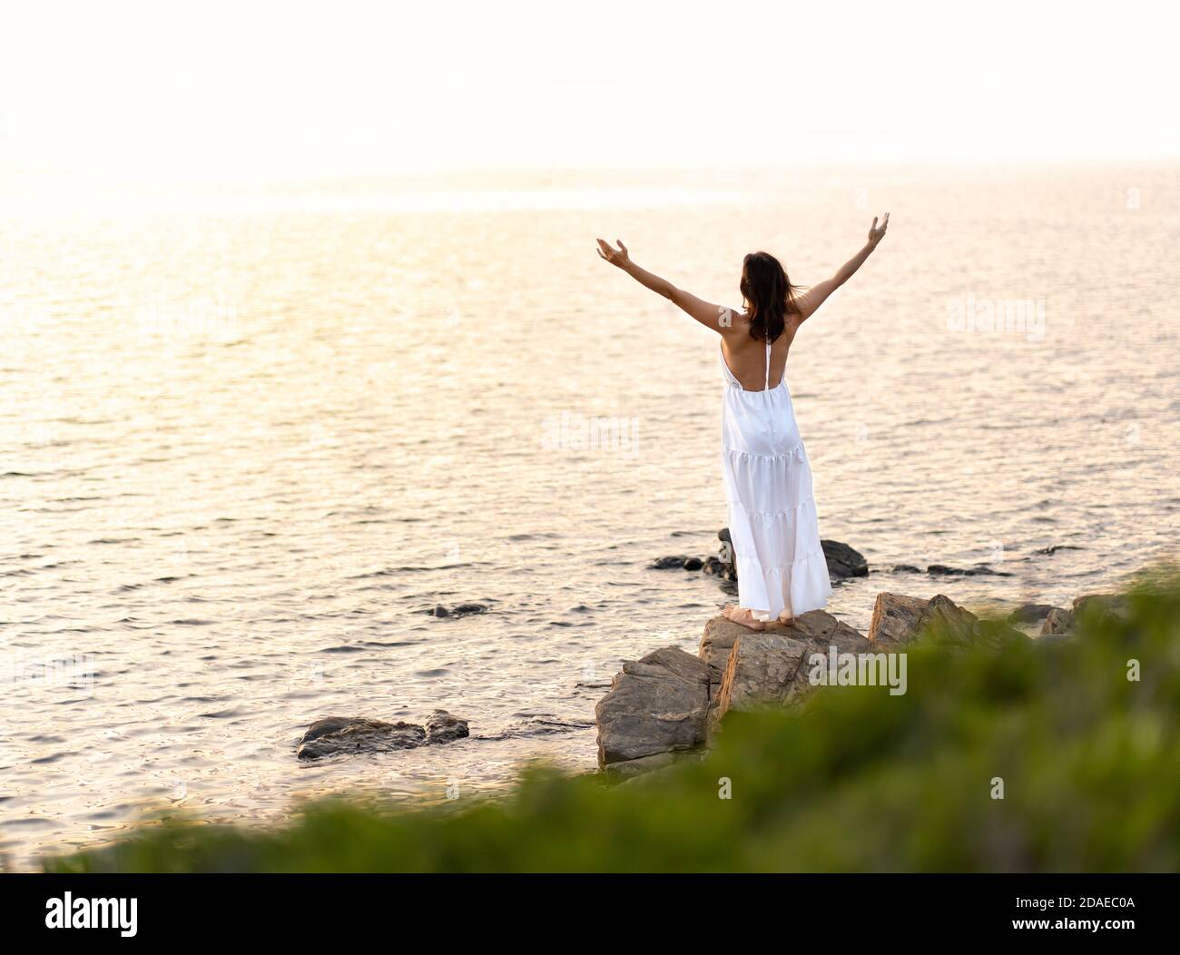 Vue de l'arrière d'une femme méconnaissable avec une longue blanche habillez-vous et ouvrez les bras vers le haut en regardant le soleil couchant - seule femme avec pieds nus o Banque D'Images