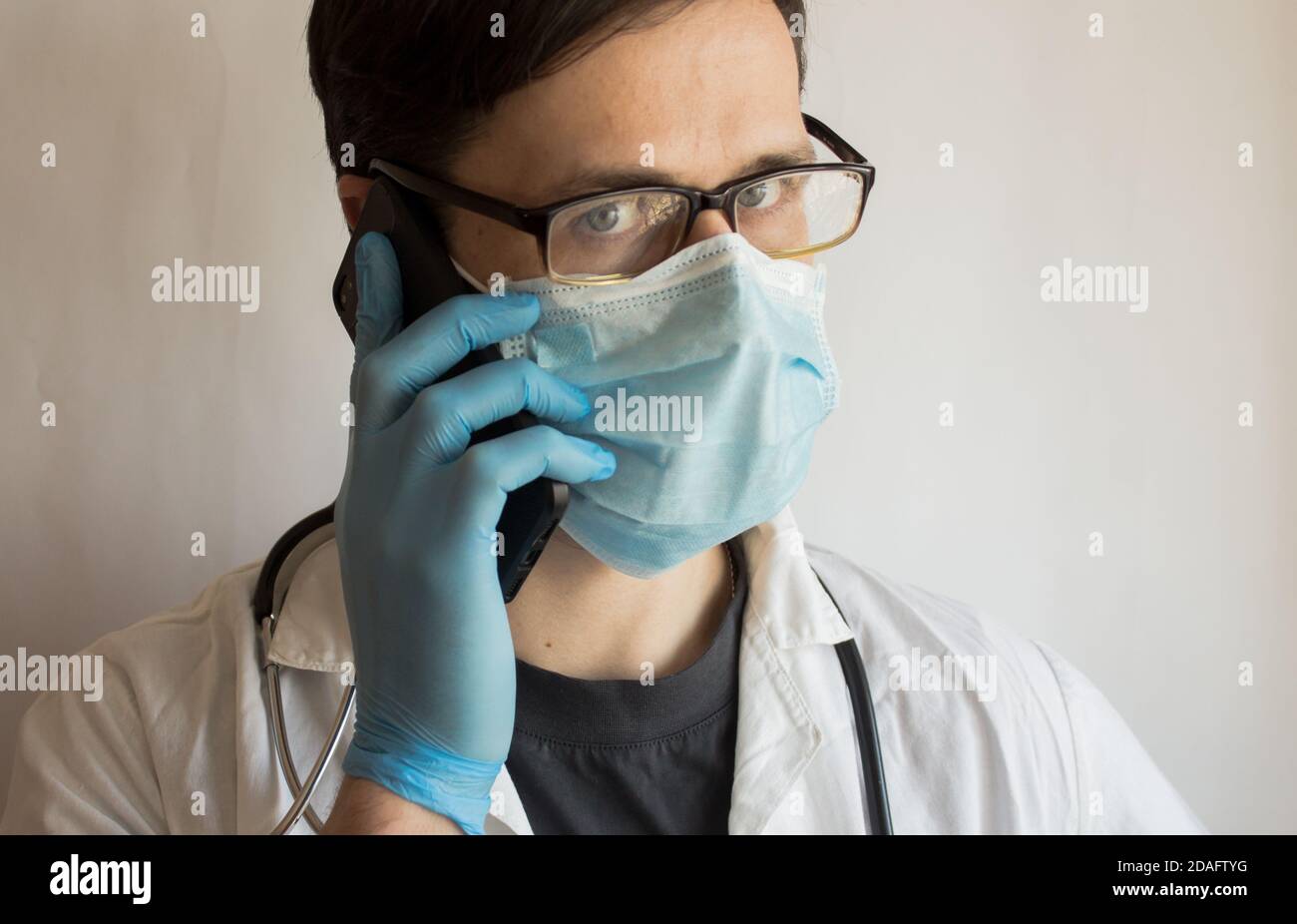 Un jeune beau médecin portant des lunettes et un masque médical donne une consultation au téléphone. Banque D'Images