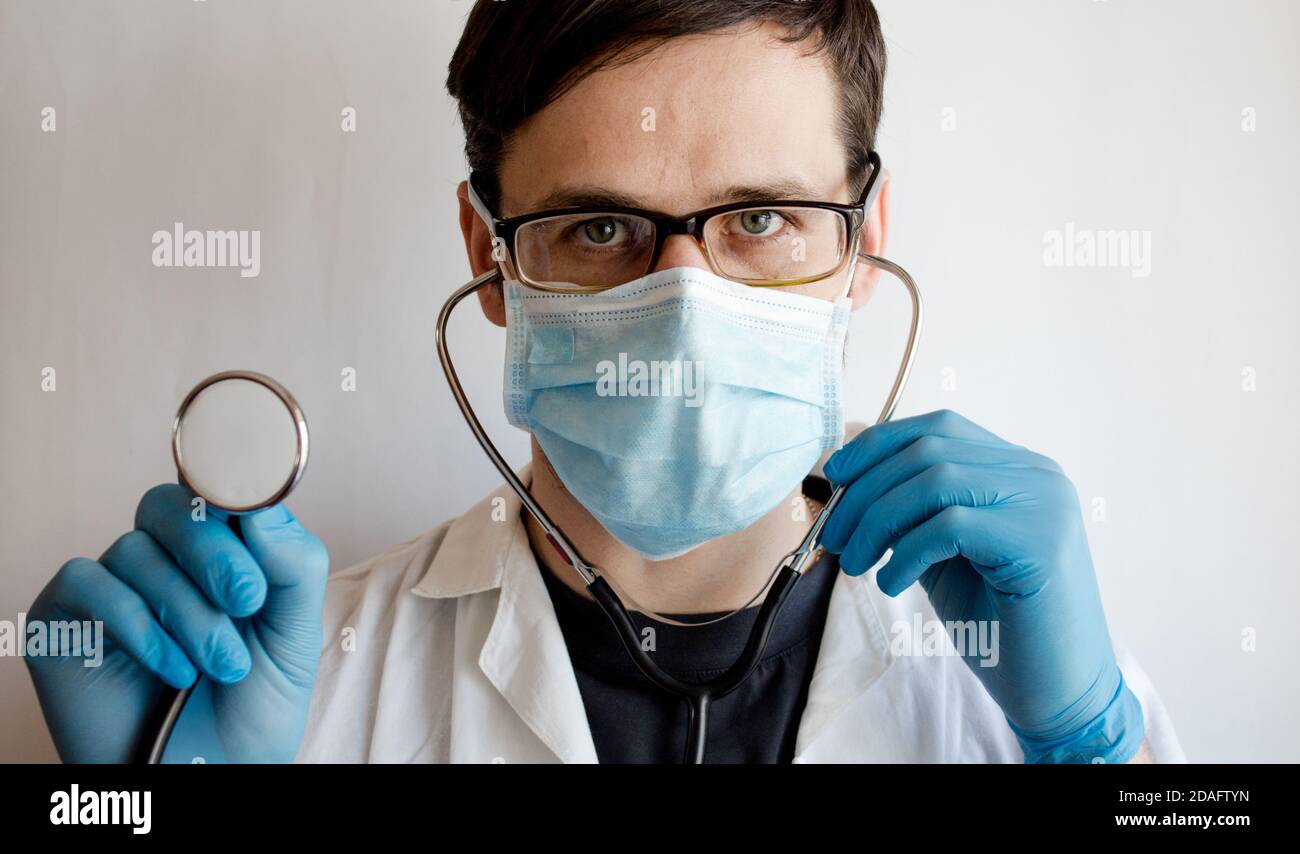 Un jeune beau médecin portant des lunettes et un masque médical met sur un stéthoscope tout en se préparant à un examen du patient. Banque D'Images