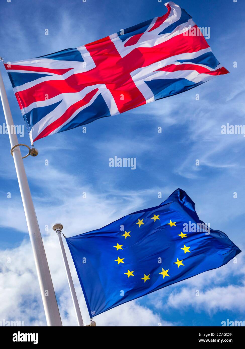 DRAPEAUX DU BREXIT CONCEPT de l'UE le drapeau britannique de l'Union Jack vole haut Au-dessus du drapeau européen de l'UE dans une brise raide sur un journée ensoleillée avec ciel bleu et drapeaux entourés de tourbillons nuages Banque D'Images