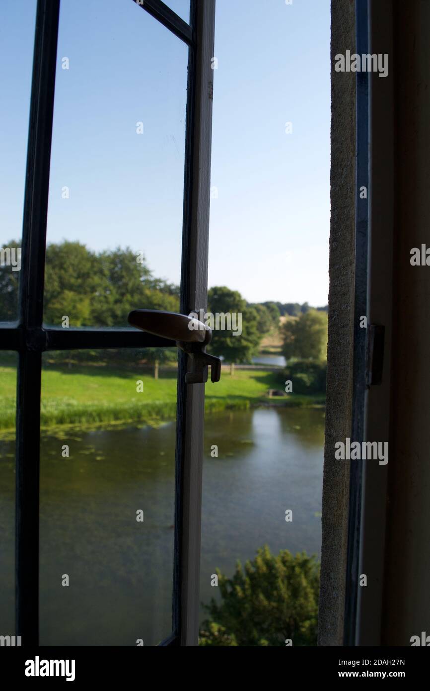 Vue à travers une fenêtre en métal partiellement ouverte sur une scène de campagne d'été : herbe verte, arbres (bois) et un lac ou une rivière tranquille Banque D'Images