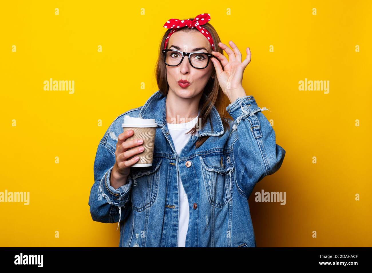 Jeune femme en veste denim, serre-tête et lunettes tenant une tasse en papier dans les mains sur fond jaune Banque D'Images