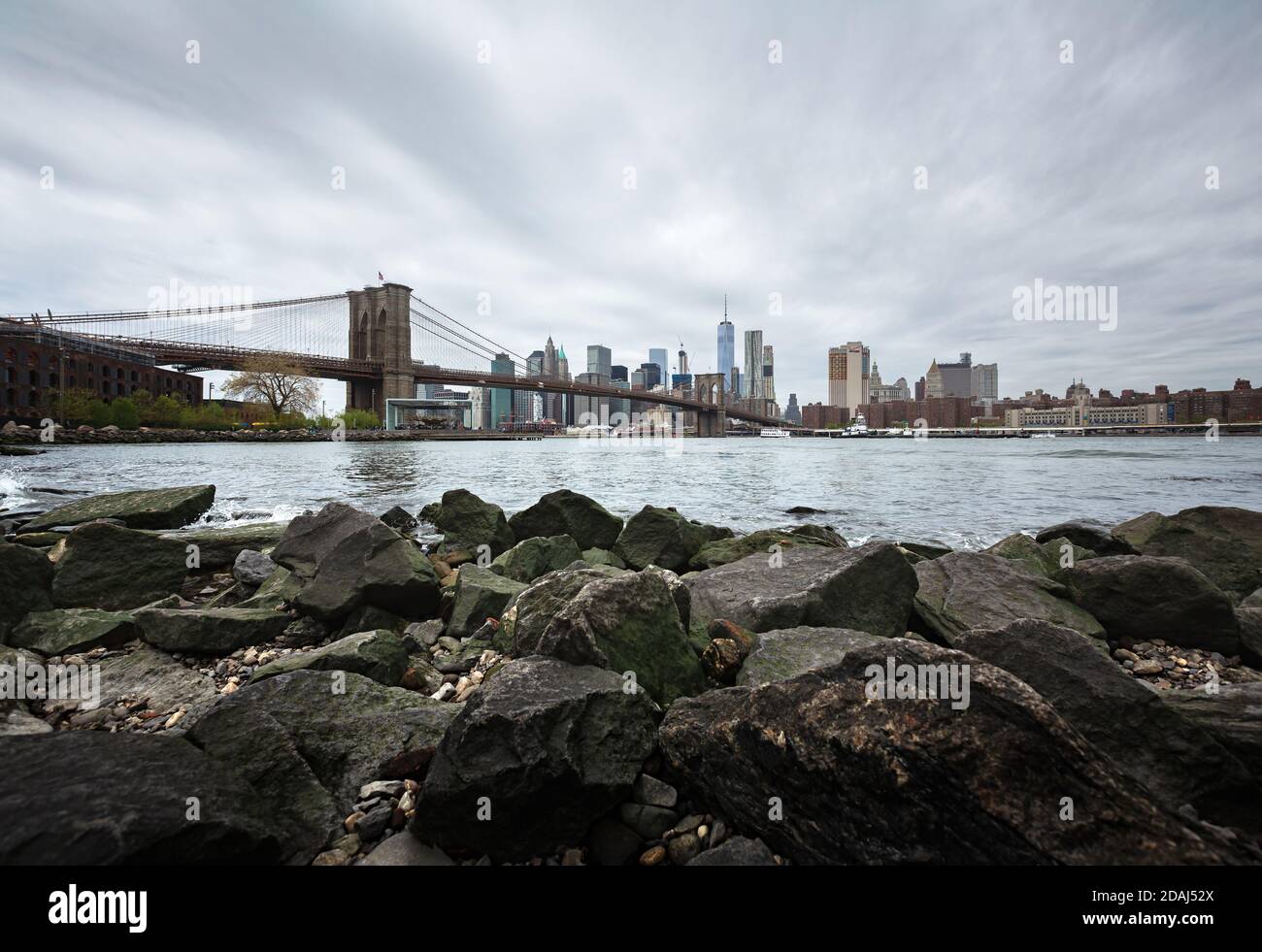 NEW YORK, États-Unis - 29 avril 2016 : gratte-ciel de Manhattan avec pont de Brooklyn. Rochers et pierres sur la rive de la rivière est. Horizon de Manhattan depuis Dumbo Banque D'Images