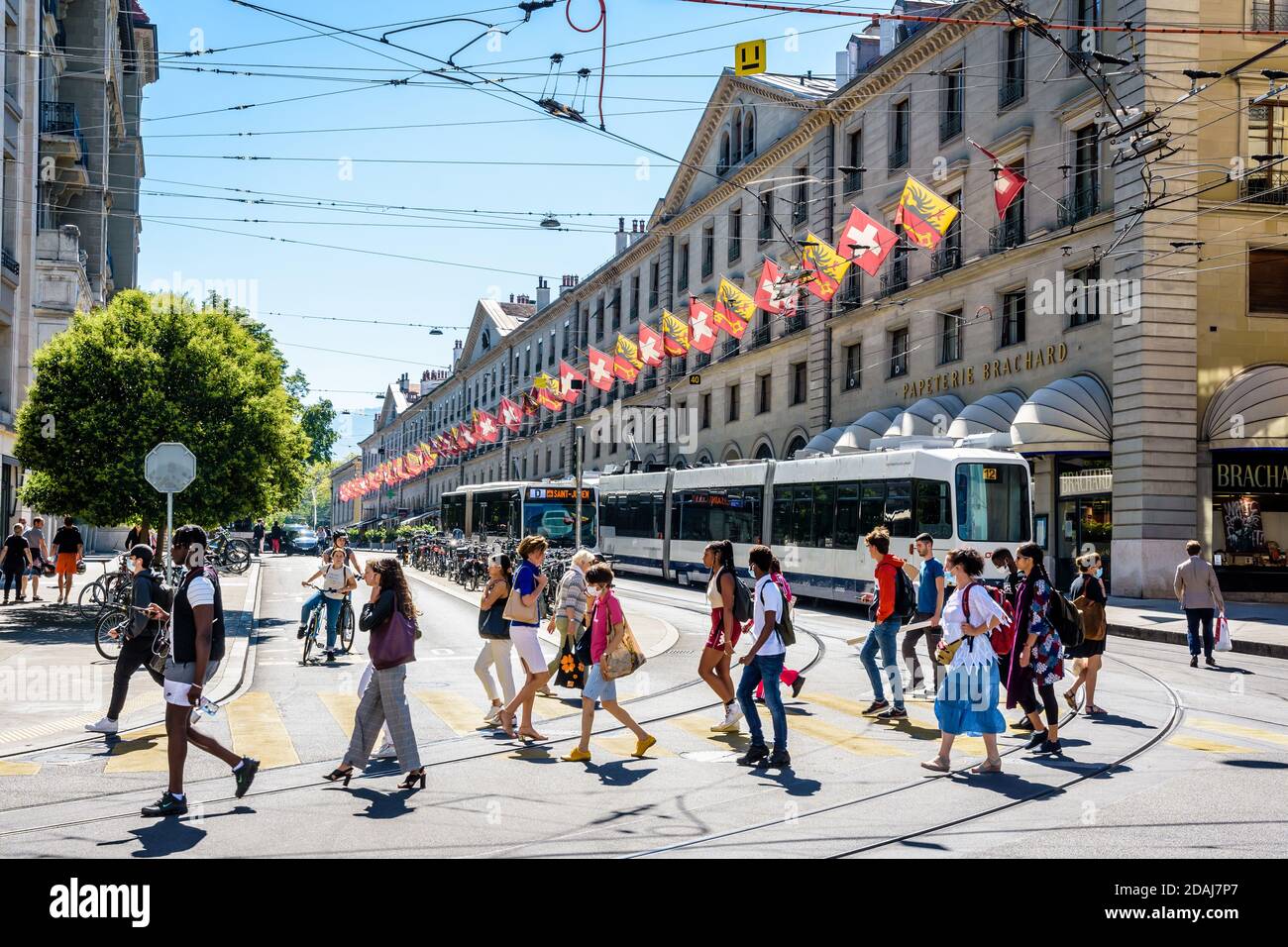 Des piétons traversent la rue de la Corraterie à Genève, avec des bâtiments avec des drapeaux, comme un bus et un tramway passent l'un l'autre. Banque D'Images