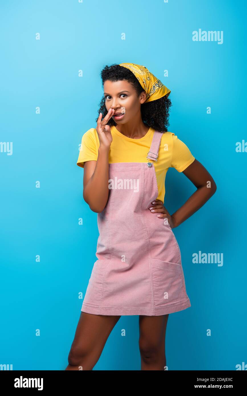 Femme afro-américaine debout avec la main sur la hanche et le gossiping sur bleu Banque D'Images