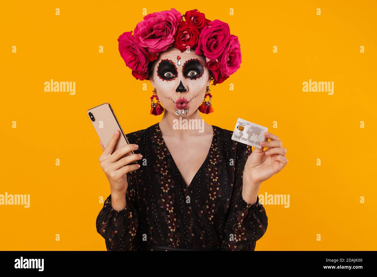 Une jeune fille choquée dans le maquillage d'halloween posant avec le téléphone cellulaire et carte de crédit isolée sur un mur jaune Banque D'Images