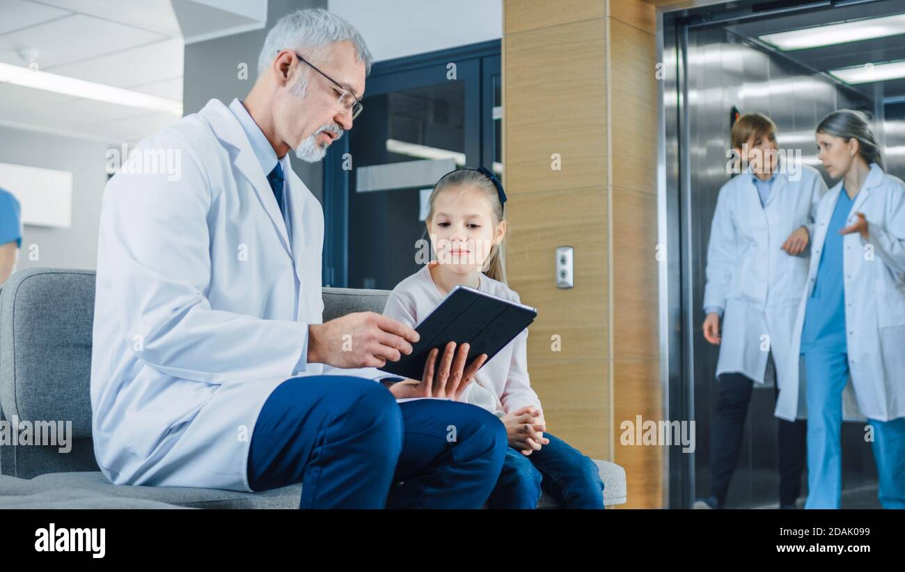 Dans le hall de l'hôpital, un médecin masculin parle avec une petite fille mignonne tout en étant assis sur le canapé, il lui montre des informations sur l'ordinateur tablette Banque D'Images