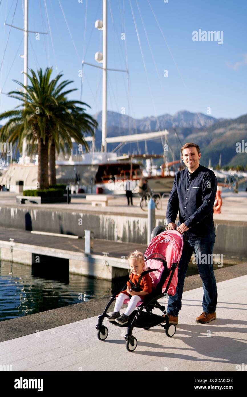 L'homme pousse une poussette rose avec sa fille à l'intérieur Sur une promenade à côté des bateaux au Monténégro Banque D'Images
