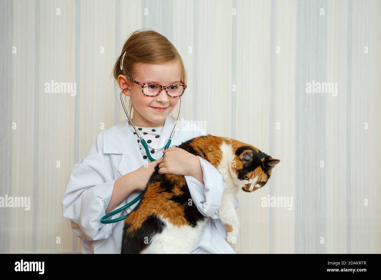 Une petite fille dans un manteau de docteur sourit et invite à un traitement. Le patient est un chat domestique. Jeu médical éducatif. Banque D'Images