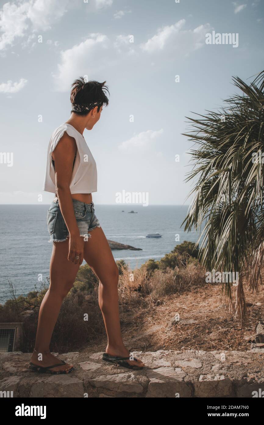 Femme attrayante avec des cheveux courts regardant la mer pendant Marche en haut d'une falaise à Ibiza avec du denim shorts Banque D'Images