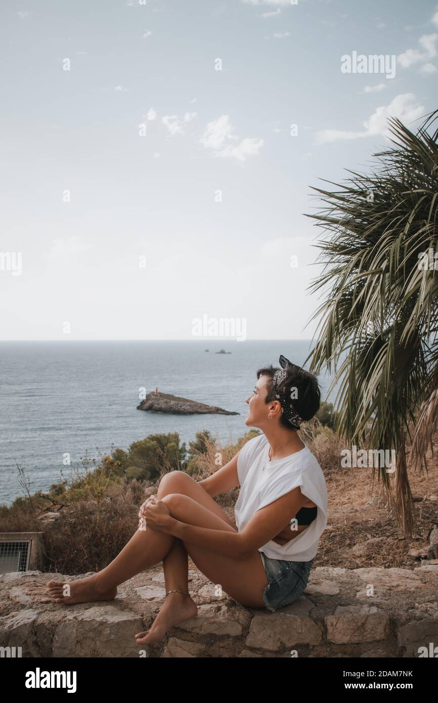 Belle femme avec des cheveux courts regardant la mer souriant Assis  au-dessus d'une falaise à Ibiza, portant un short en denim Photo Stock -  Alamy