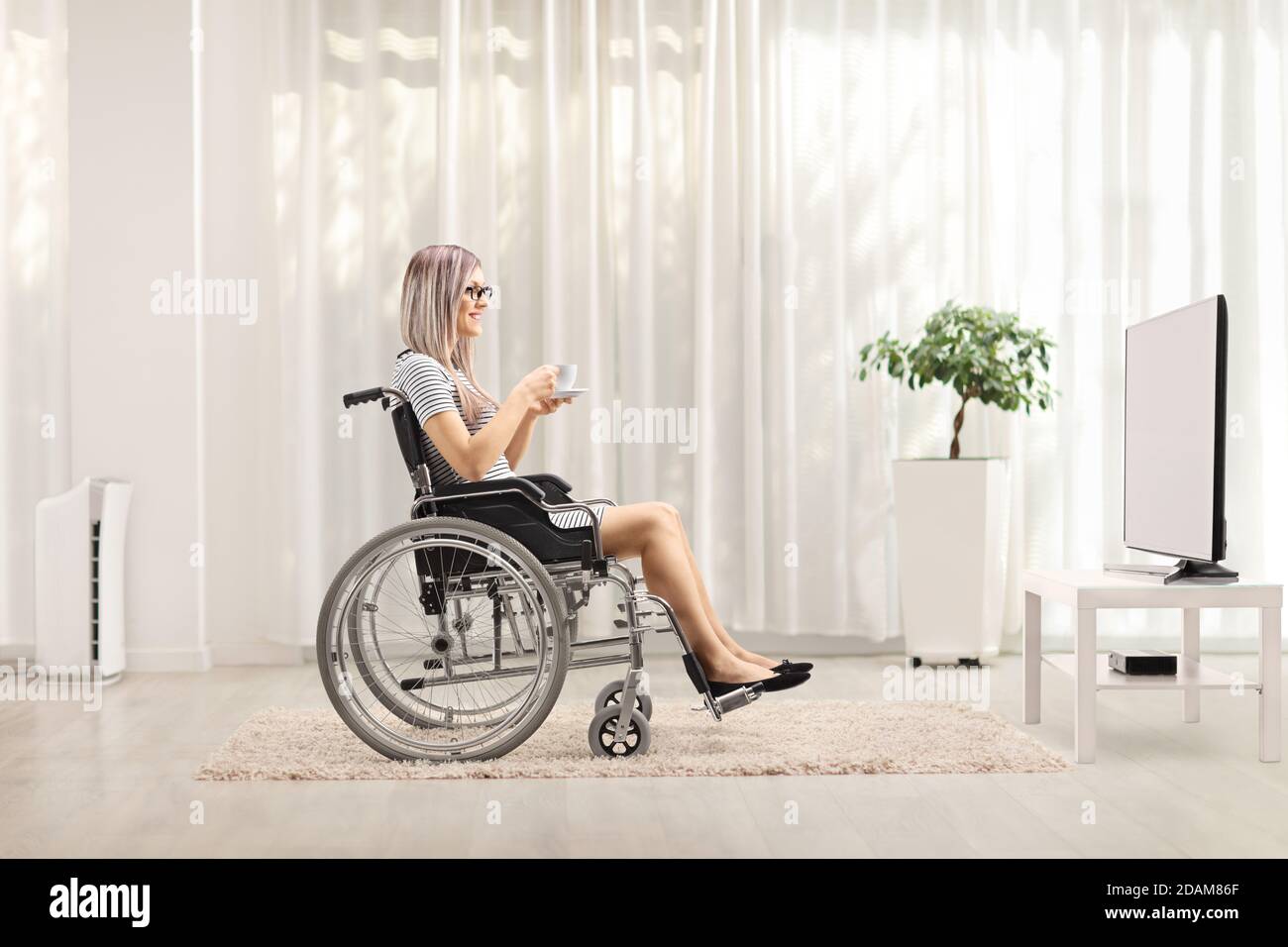 Photo de profil d'une jeune femme en fauteuil roulant avec une tasse de thé devant la télévision à la maison Banque D'Images