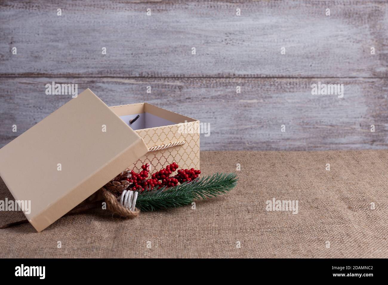 boîte cadeau beige carte postale avec couvercle ouvert et branche De sapin de Noël avec des décorations sur fond de bois et de toile de jute Banque D'Images