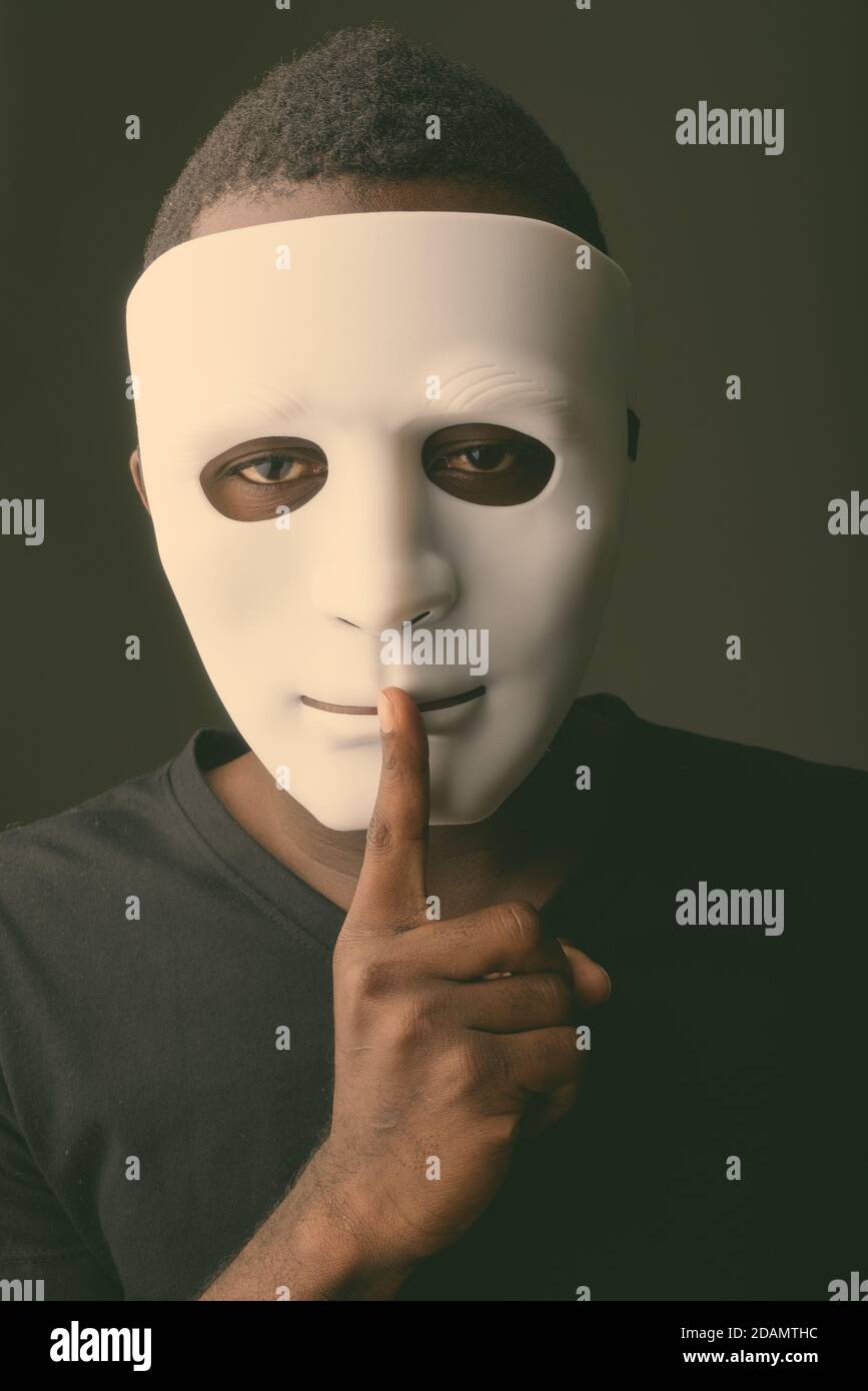 Studio de photo d'un jeune homme noir africain dans une pièce sombre porter un masque et tenir le doigt sur les lèvres Banque D'Images