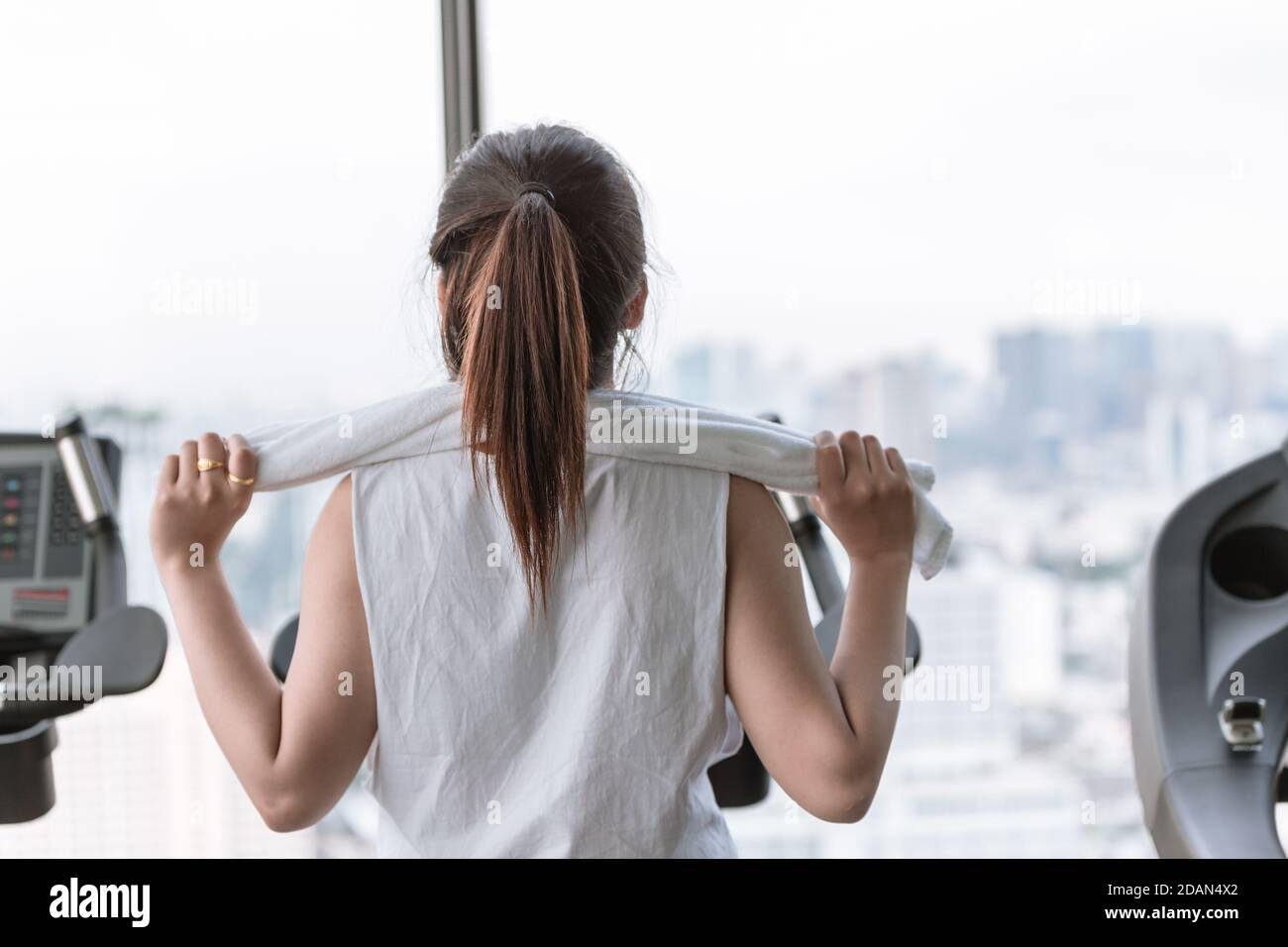 Femme asiatique exercice matinal dans le club de sport avec une serviette sur ses épaules en regardant la ville aux fenêtres vue de derrière. Banque D'Images