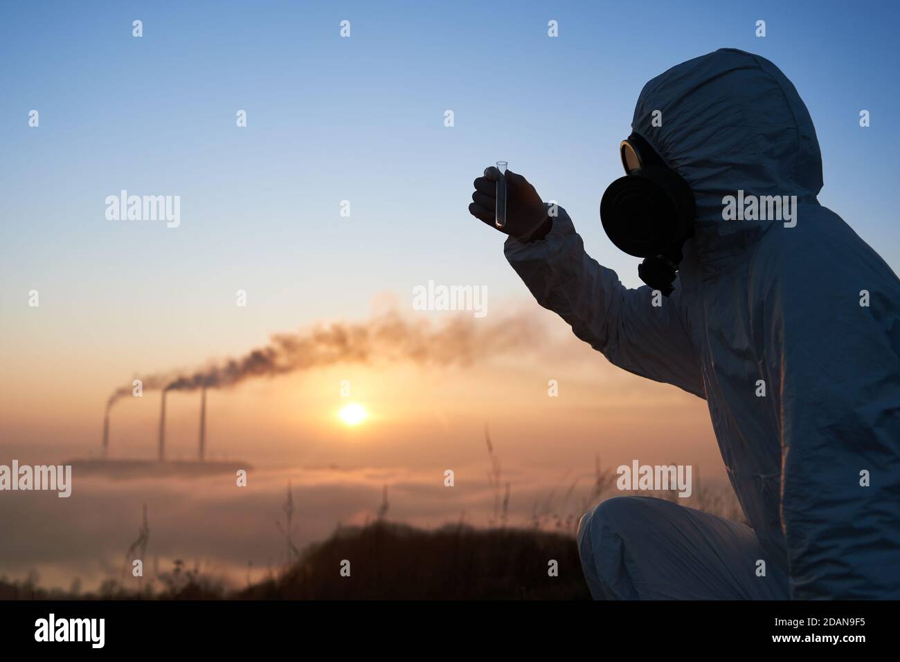 Gros plan de l'écologiste mâle dans le masque à gaz contenant le tube à essai avec l'échantillon de sol. Scientifique explorant les questions environnementales à la centrale électrique avec des cheminées de fumée et un ciel magnifique en arrière-plan. Banque D'Images