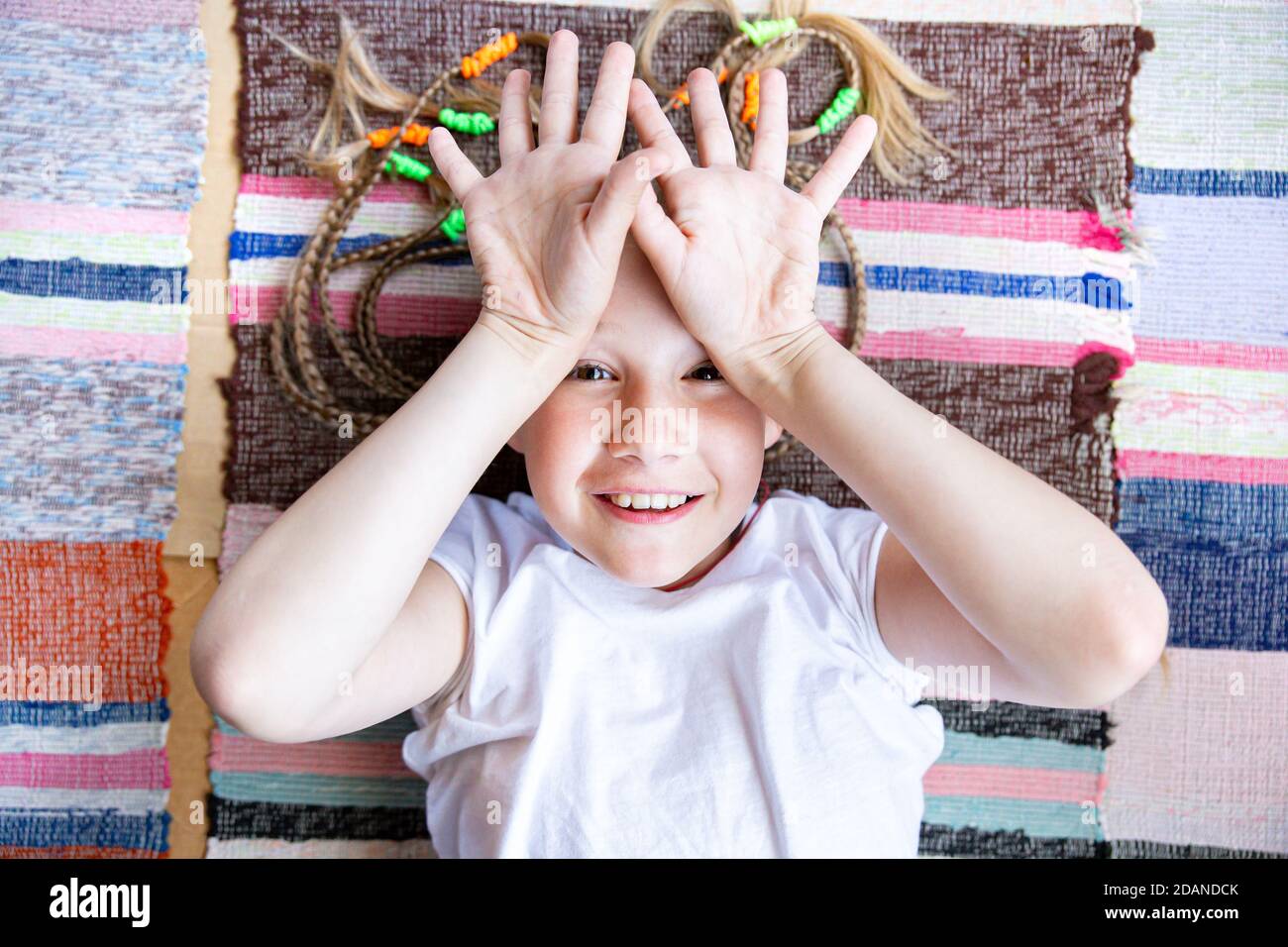 Un enfant caucasien, une fille gaie avec des picots, se trouve sur le sol sur un tapis tissé rayé, sourires, montre des palmiers ouverts avec les doigts écartés. Gros plan sur la Banque D'Images