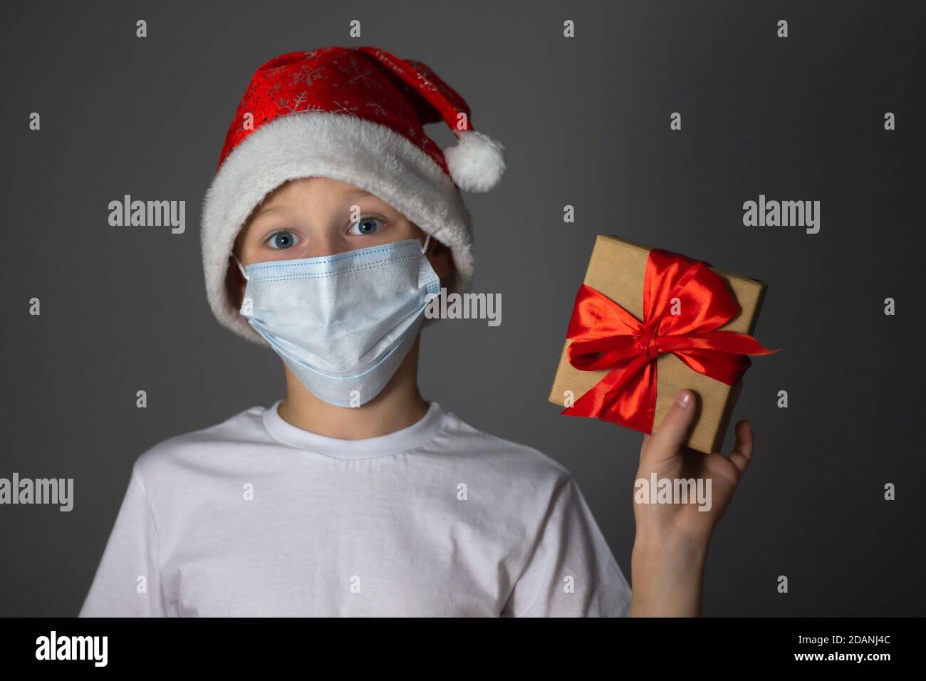 Un garçon de 7-10 ans dans un T-shirt blanc, une casquette de Noël et un masque médical tenant un cadeau sur fond gris. Noël et nouvel an pendant la pandémie du coronavirus Banque D'Images