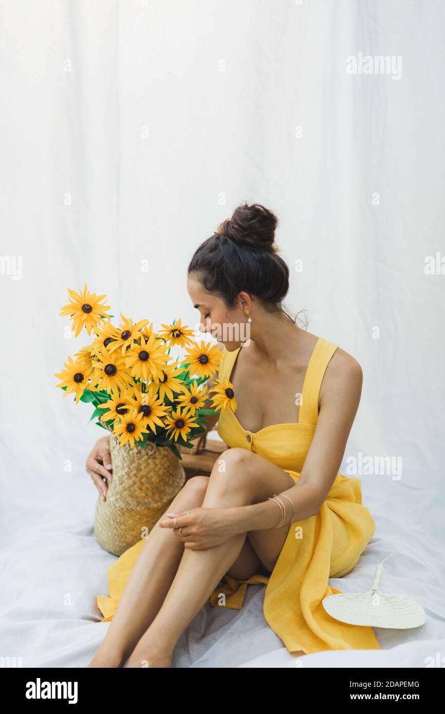 Portrait de jeune femme brune sur fond de tissu blanc suspendu avec panier de paille de fleurs jaunes, robe de lin d'été. Banque D'Images