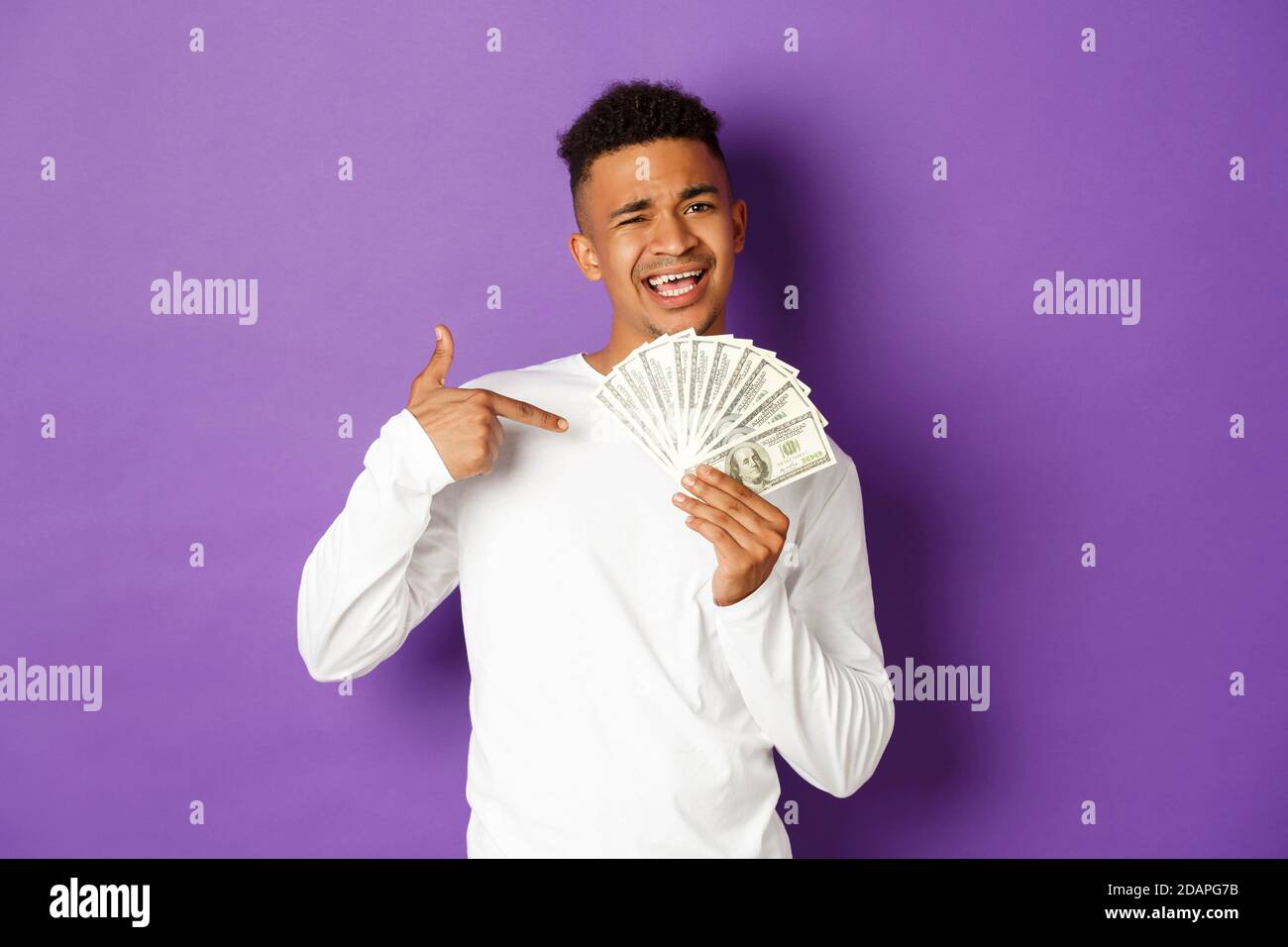 Image d'un homme afro-américain assité souriant, se vanchant avec une grosse somme d'argent, pointant de l'argent, se tenant sur fond violet Banque D'Images