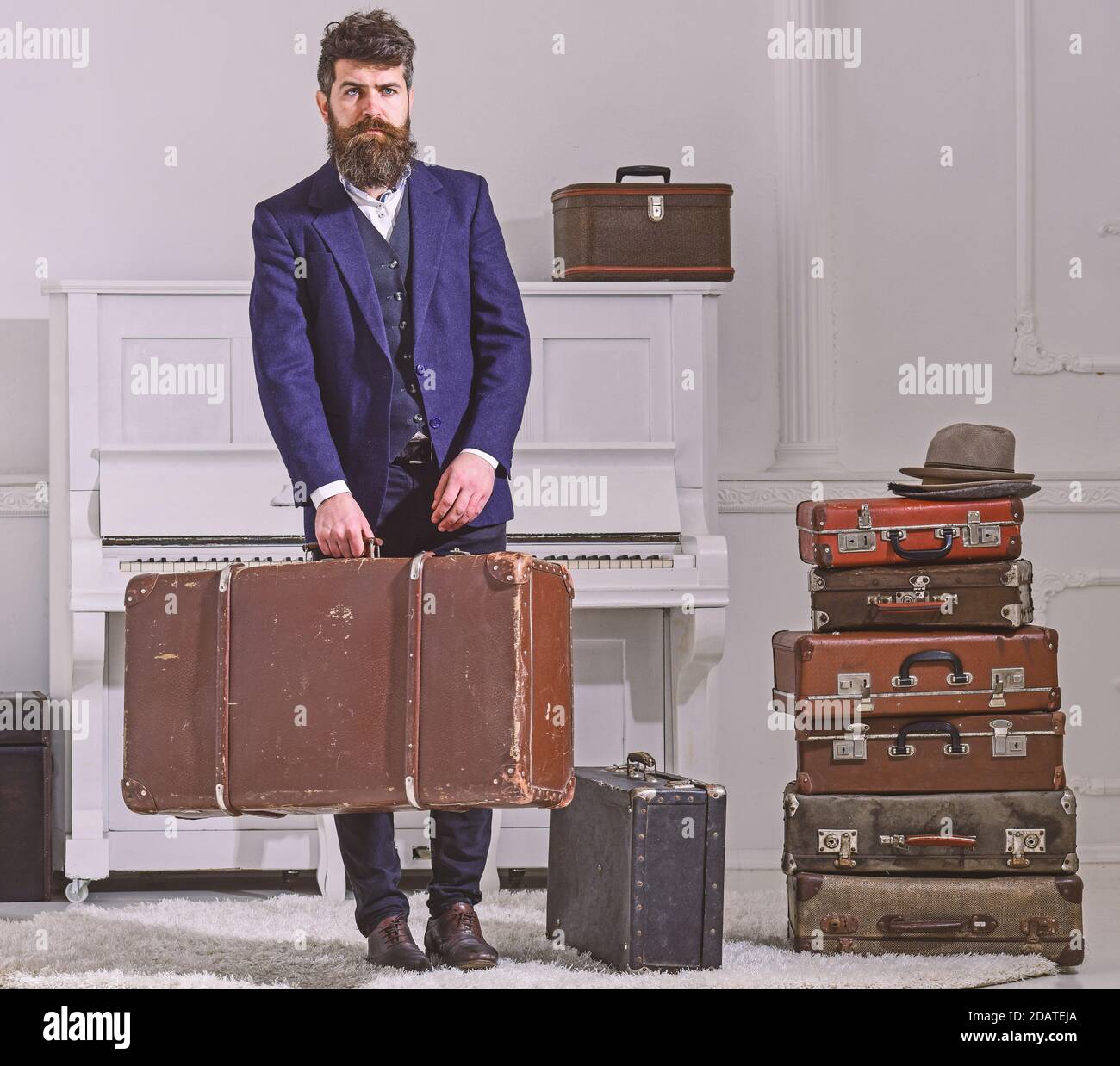 Macho élégant sur le visage strict se tient près de pile de valise vintage,  tient la valise. Homme, voyageur avec barbe et moustache avec bagages,  arrière-plan intérieur blanc de luxe. Concept de