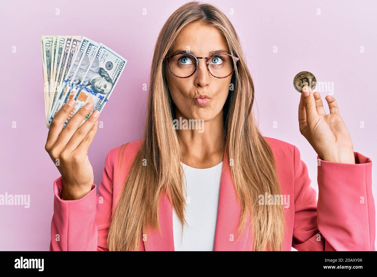 Jeune femme blonde portant le style d'affaires tenant des dollars et bitcoin faire le visage de poisson avec la bouche et les yeux de accroupi, fou et comique. Banque D'Images