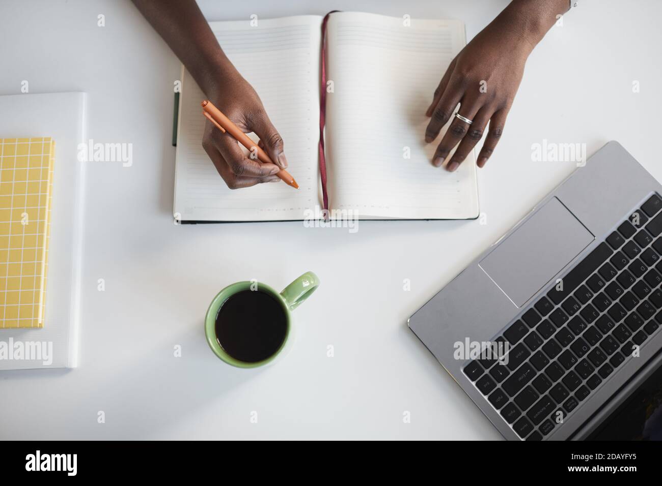 Vue minimale sur le dessus d'une femme afro-américaine méconnaissable qui écrit dans un planificateur sur un lieu de travail blanc Banque D'Images