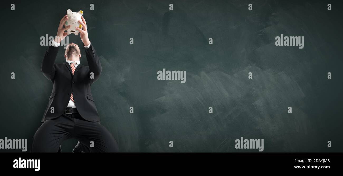 un homme d'affaires kneeing tenant une tirelire au-dessus de sa tête devant lui d'un tableau noir Banque D'Images