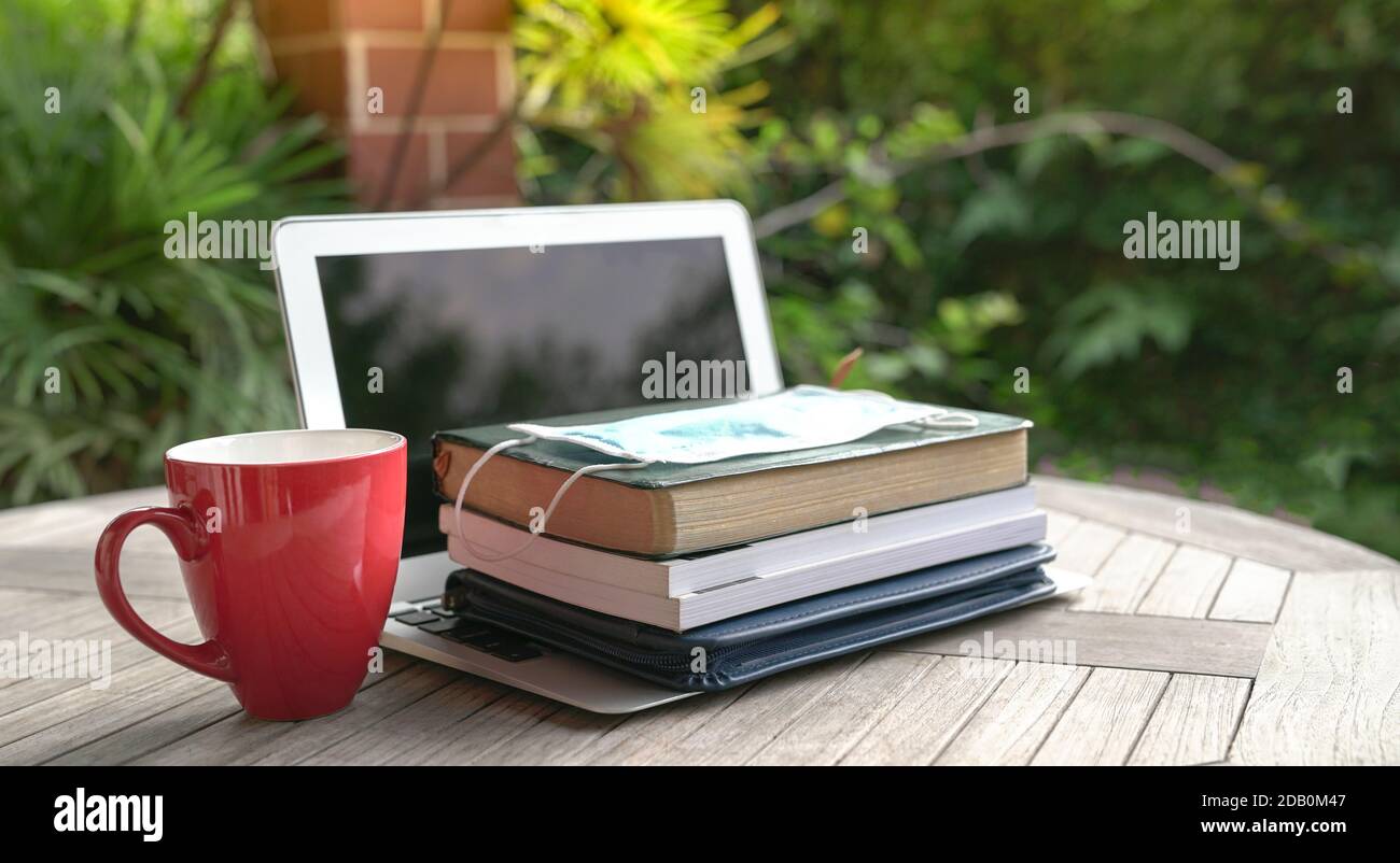 Masque sur le dessus des livres empilés et ordinateur portable ouvert avec une tasse rouge. Étudier à domicile pendant le concept de pandémie. Banque D'Images