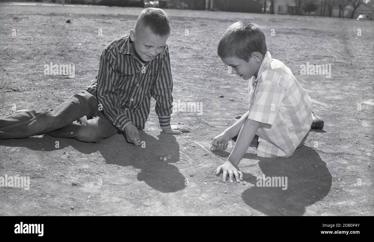 Années 1960, historique, deux jeunes garçons à l'extérieur dans une zone de gravier jouant un jeu de billes, USA. Banque D'Images