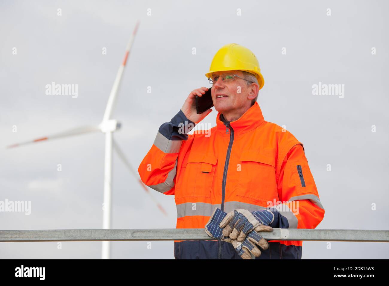 Ingénieur portant un vêtement de protection pour parler sur un téléphone portable avant d'une éolienne en arrière-plan flou Banque D'Images