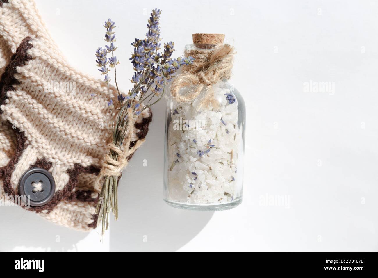 Une bouteille de sel de mer en verre pour un bain relaxant sur fond blanc à côté d'un petit bouquet de lavande et avec un détail en maille beige et un brun Banque D'Images