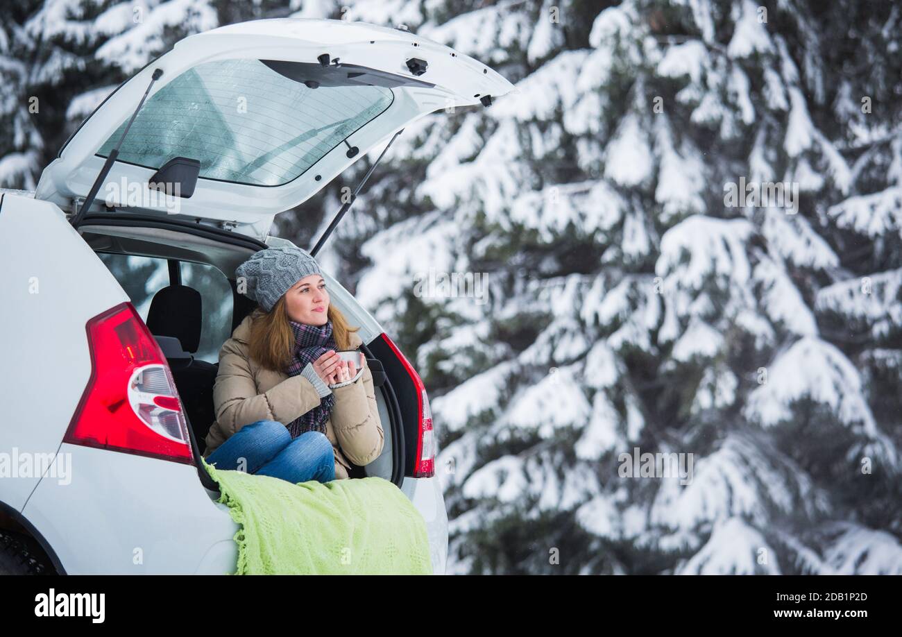 La femme est assise dans le coffre de la voiture et tient une tasse de thé chaud dans ses mains. Banque D'Images