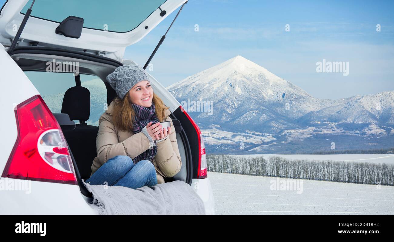 La femme est assise dans le coffre de la voiture et tient une tasse de thé chaud dans ses mains. Banque D'Images