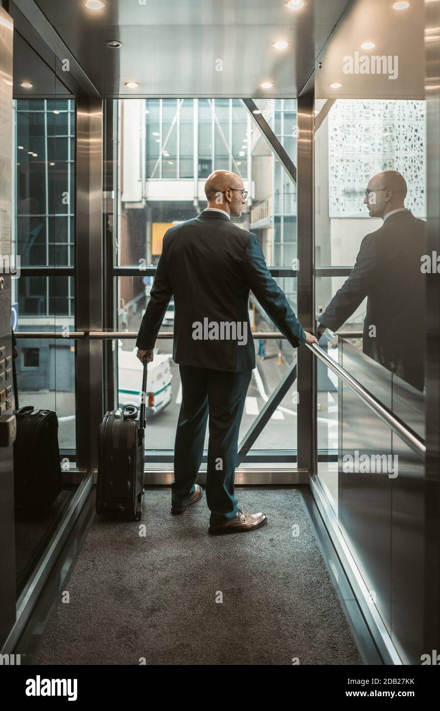 Homme d'affaires avec valise dans le centre d'affaires. Concept de voyage d'affaires. Un homme de race blanche d'âge moyen regarde par la paroi en verre de l'ascenseur. Vue arrière Banque D'Images