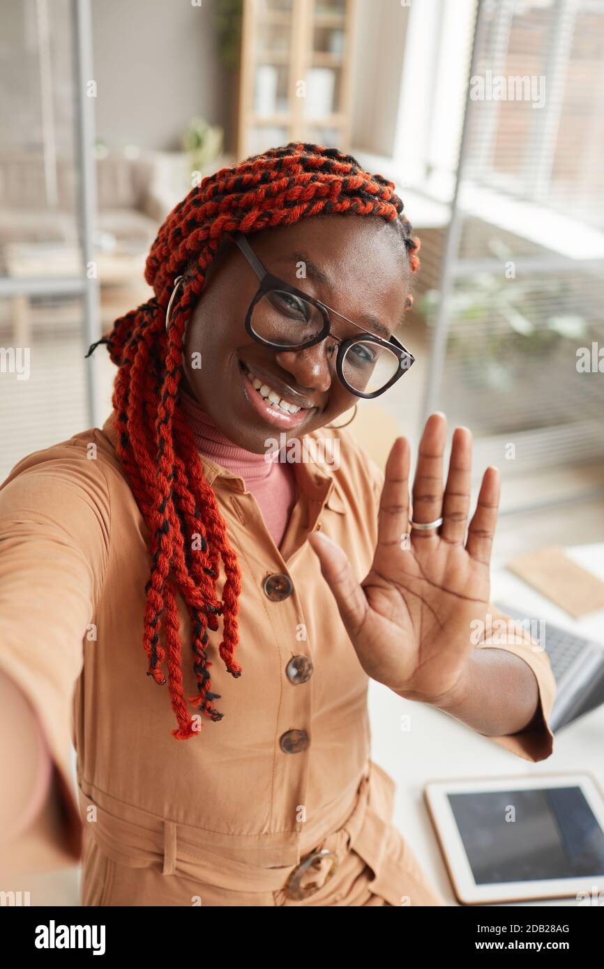 Portrait grand angle de la jeune femme afro-américaine qui agite à la caméra lors de la prise de photos de selfie ou de la diffusion en direct à partir du bureau à domicile Banque D'Images