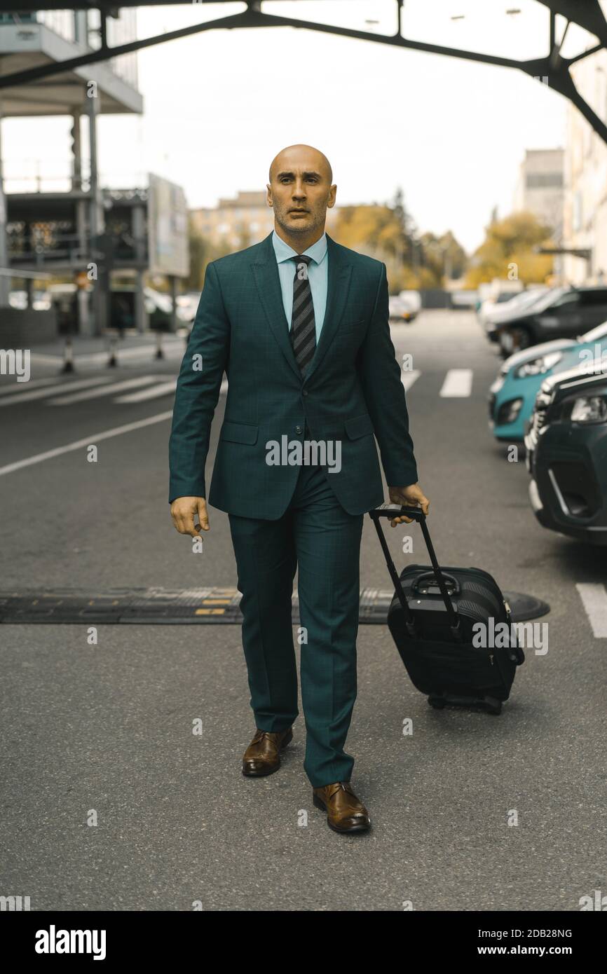 L'homme d'affaires va de l'avant en tenant sa valise en main. Un bel homme du Caucase qui passe devant les voitures garées de l'hôtel. Concept de voyage d'affaires Banque D'Images