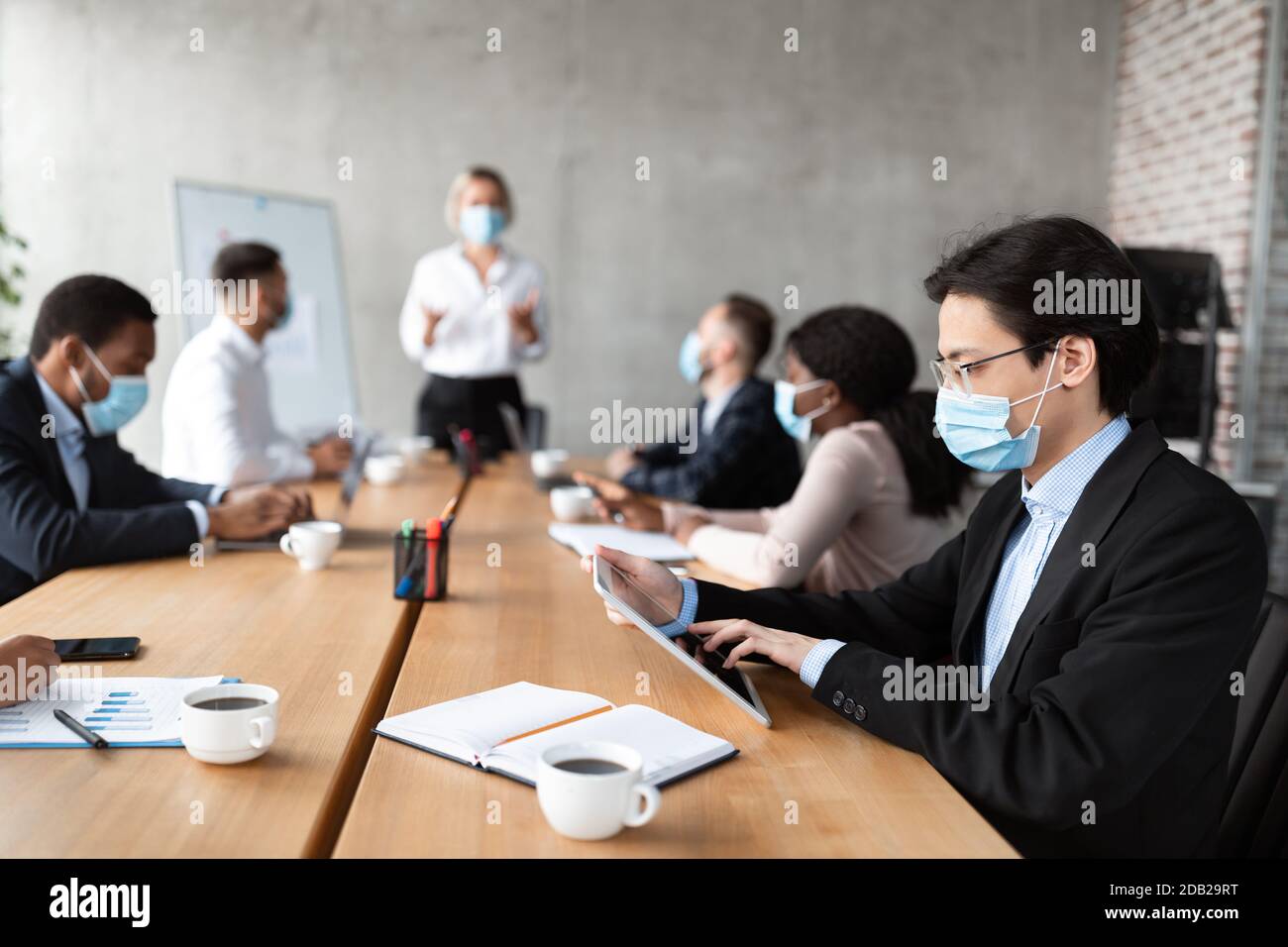 Homme d'affaires asiatique en masque utilisant une tablette pendant une réunion d'entreprise en intérieur Banque D'Images