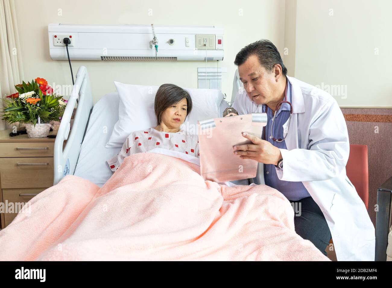 Médecin asiatique confiant explantant à une patiente gaie sur un état de santé allongé sur le lit à l'hôpital. Banque D'Images
