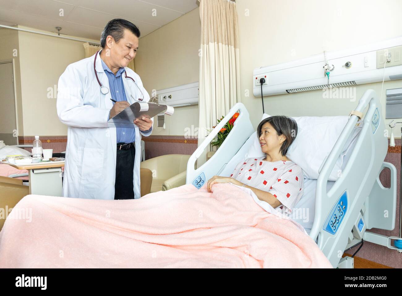 Médecin asiatique confiant explantant à une patiente gaie sur un état de santé allongé sur le lit à l'hôpital. Banque D'Images