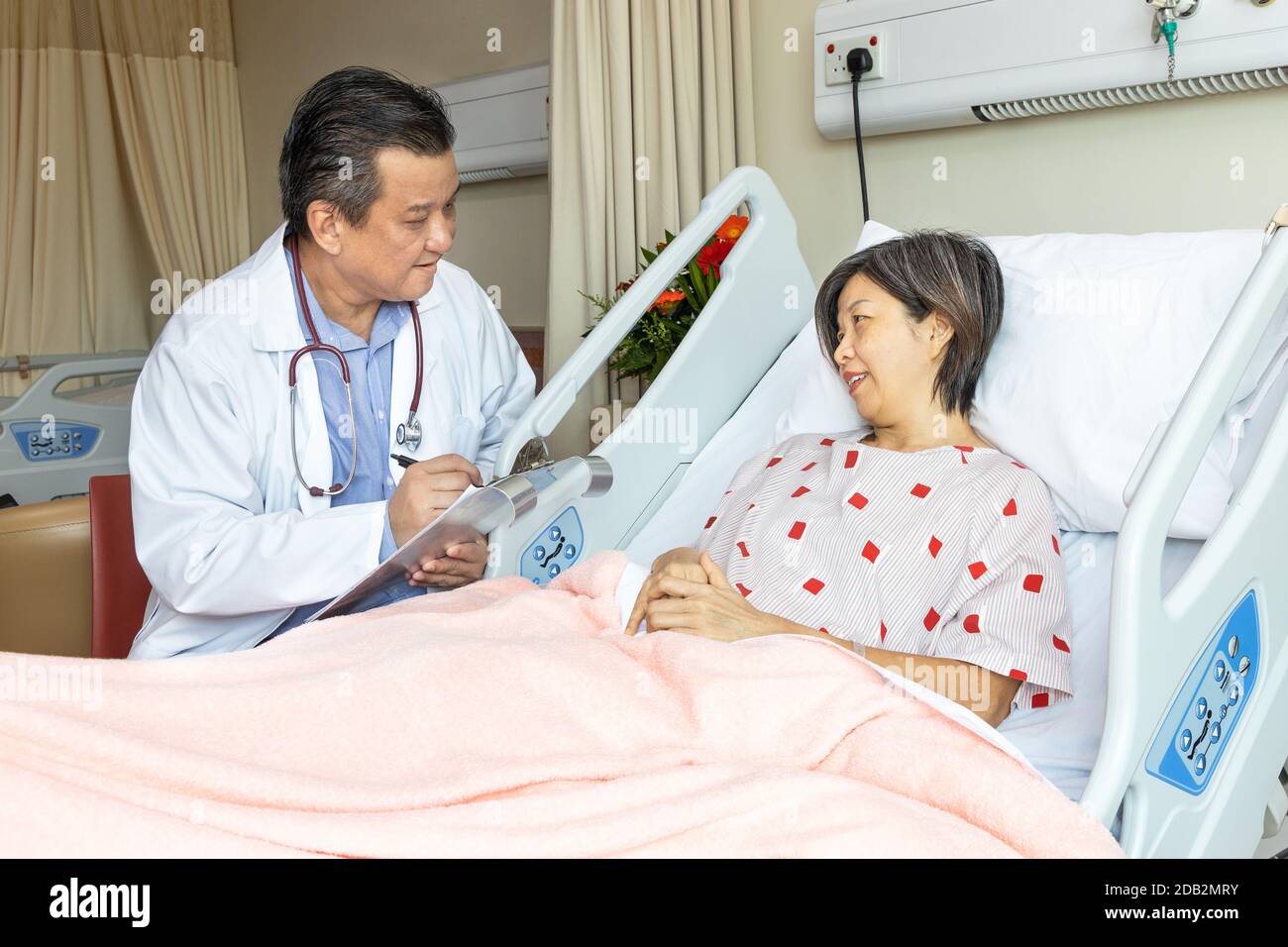 Médecin asiatique confiant explantant à une patiente gaie sur un état de santé allongé sur le lit à l'hôpital. Banque D'Images