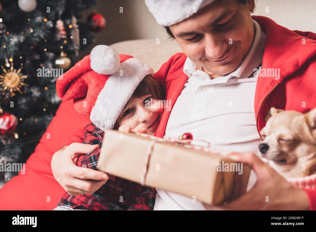 Bonne famille en chapeau de père Noël, père et enfant fils donnant cadeau de Noël à la maison. Assis sur un canapé dans la salle de séjour avec le chiot en chandail Banque D'Images