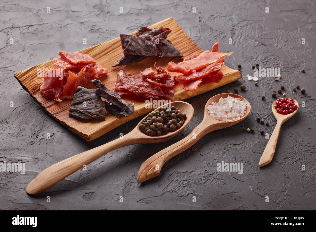 Saccadé. Ensemble de divers types de viande épicée séchée sur plateau en bois, divers grains de poivre et sel sur des cuillères en bois sur fond gris foncé. En-cas pour la bière Banque D'Images
