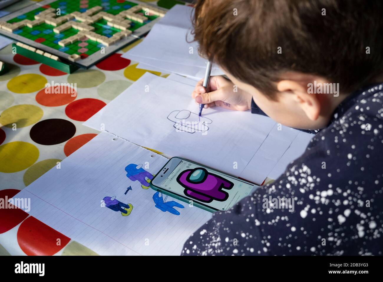 Garçon 11 enfant assis à la table de dessin à l'aide d'un stylo de couleur Pour dessiner des personnages du jeu vidéo « parmi nous » sur Écran iphone du téléphone portable au Royaume-Uni Banque D'Images