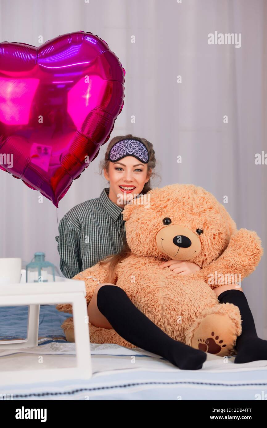 Une fille dans un masque de sommeil sourit et embrasse un ours en peluche le jour de la Saint-Valentin, à côté d'un coeur de ballon rose Banque D'Images