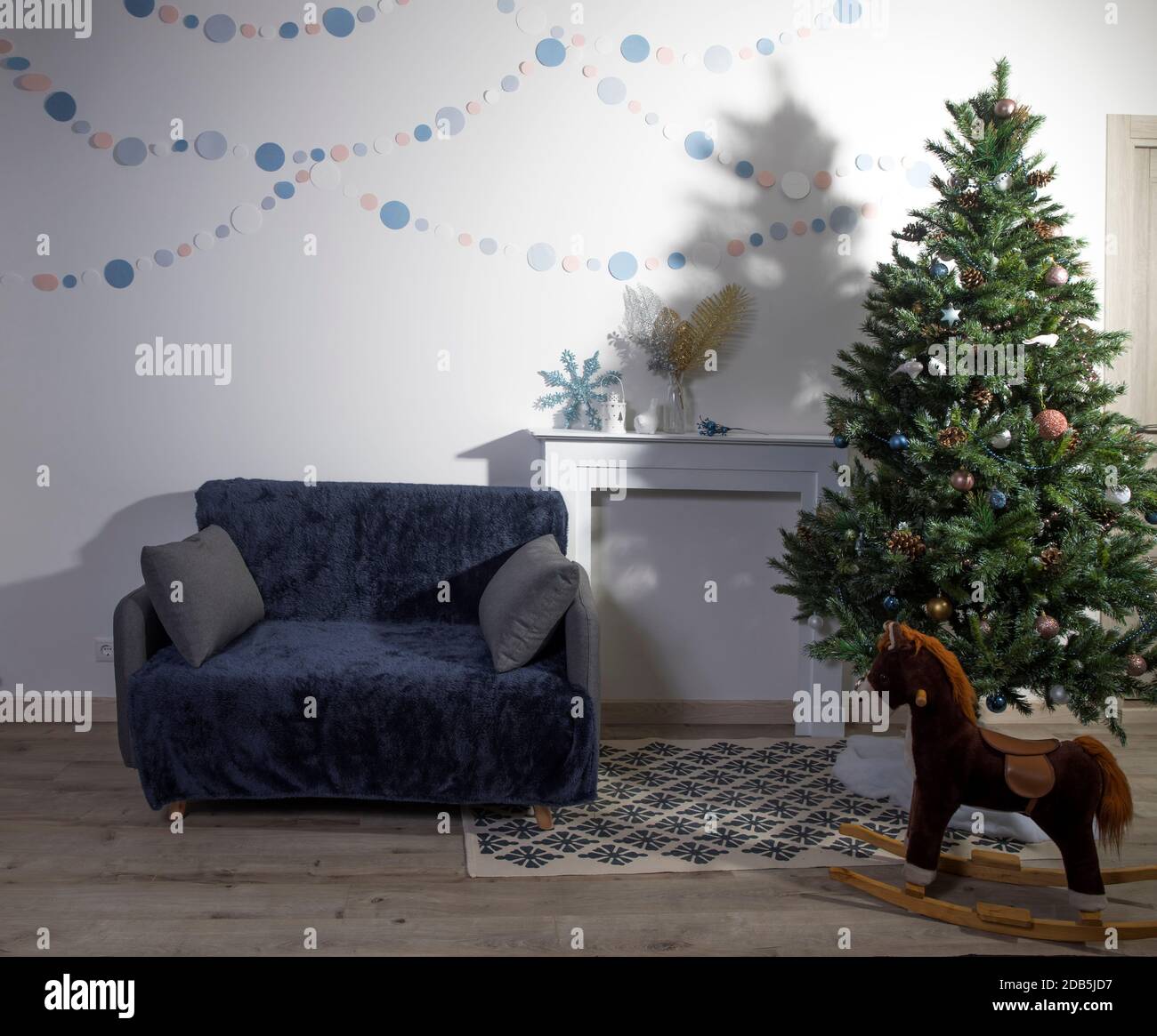 Décoration intérieure de Noël aux couleurs scandinaves blanches, un sapin de Noël décoré près d'une fausse cheminée, un cheval à bascule et un canapé. Papier garlan Banque D'Images