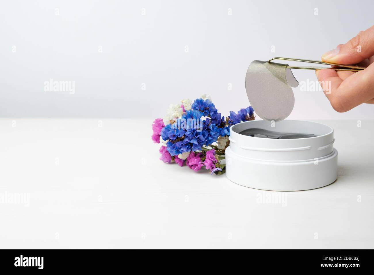 Gros plan main femme tenant un timbre d'oeil doré avec des pinces sur le fond blanc et des fleurs colorées avec espace de copie. Banque D'Images