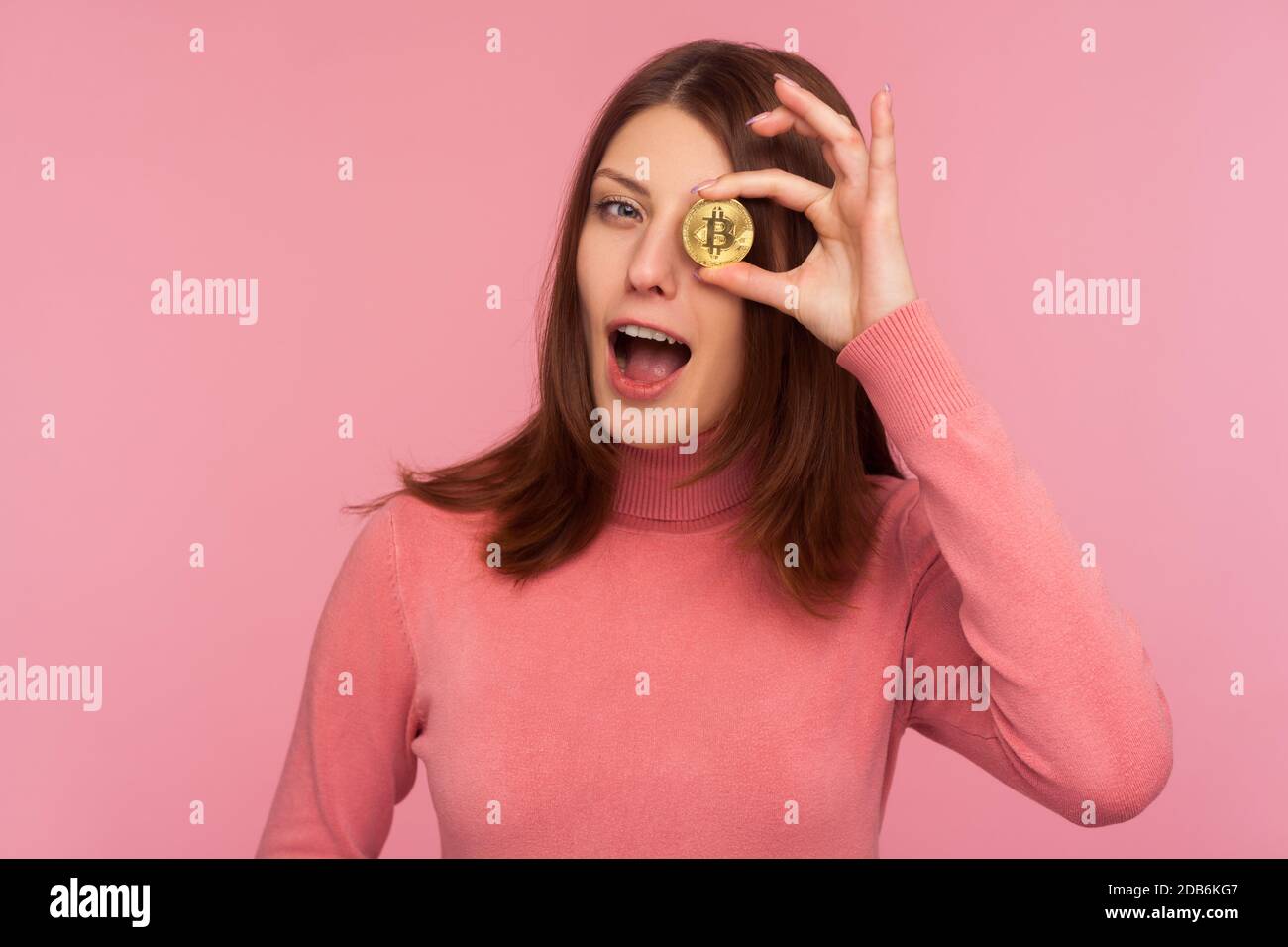 Heureuse gaie brune femme dans le chandail rose ayant l'amusement tenant la pièce de crypto près de son oeil et en se faisant un clin d'œil regardant la caméra, bitcoin crypto-monnaie. Indoo Banque D'Images