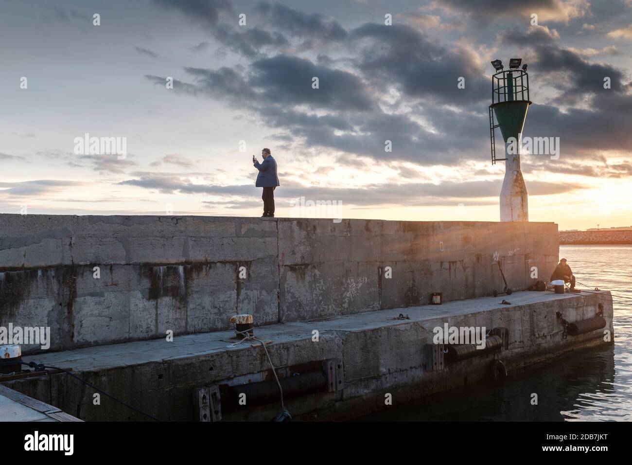 Homme d'âge moyen portant un masque facial, prenant des photos de selfie sur la jetée du port pendant le coucher du soleil, Port de Burgas, Bulgarie Banque D'Images