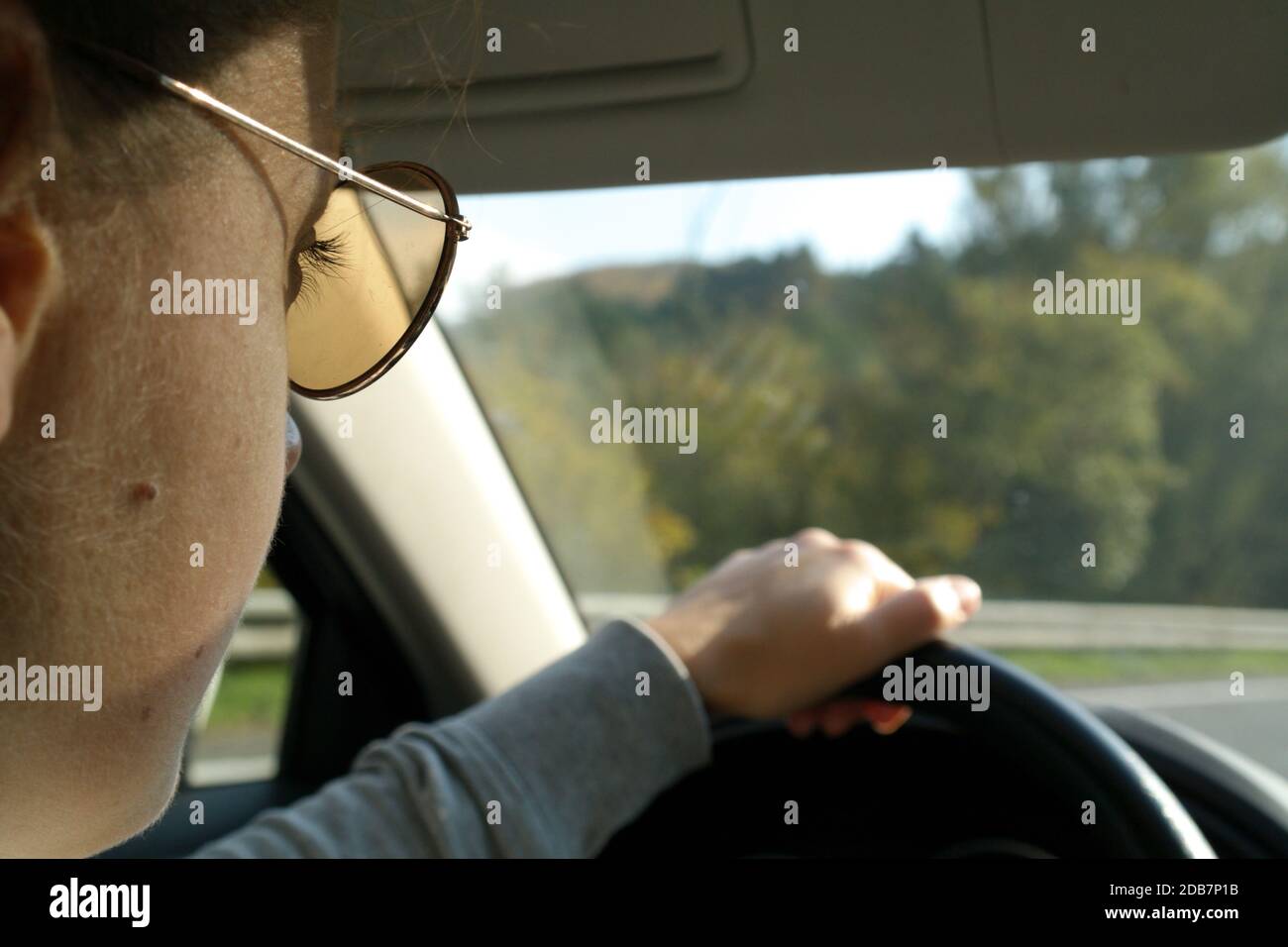 Jeune femme en lunettes de soleil conduisant une voiture sur le chemin de travailler une belle journée ensoleillée. Détail d'un côté. Roue de maintien d'une main. Banque D'Images